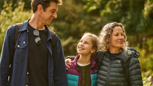 A smiling girl in a colourful padded jacket walks between two adults wearing outdoor clothing and backpacks, surrounded by sunlit trees at Lanhydrock